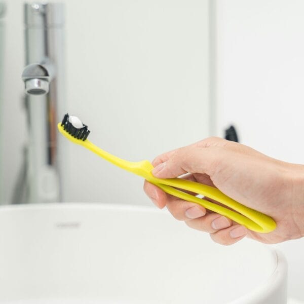 megasmile Loop toothbrush with activated charcoal bristles. Picture toothbrush yellow in the hand, over the sink. 